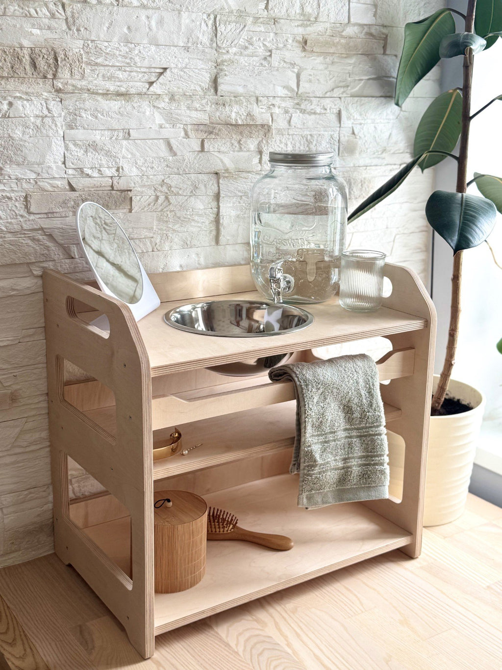 1. Montessori wooden washbasin with storage, featuring a steel bowl, towel, and accessories, set against a stone wall with a plant