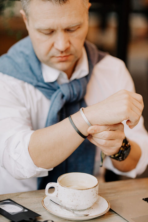 1. Man wearing a white shirt and blue sweater adjusting a silver and black bracelet at a cafe table with a coffee cup