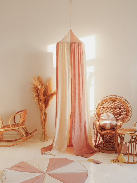 1. Powder pink and cream canopy hanging in a cozy room with wicker furniture and dried plants, creating a warm and inviting atmosphere.