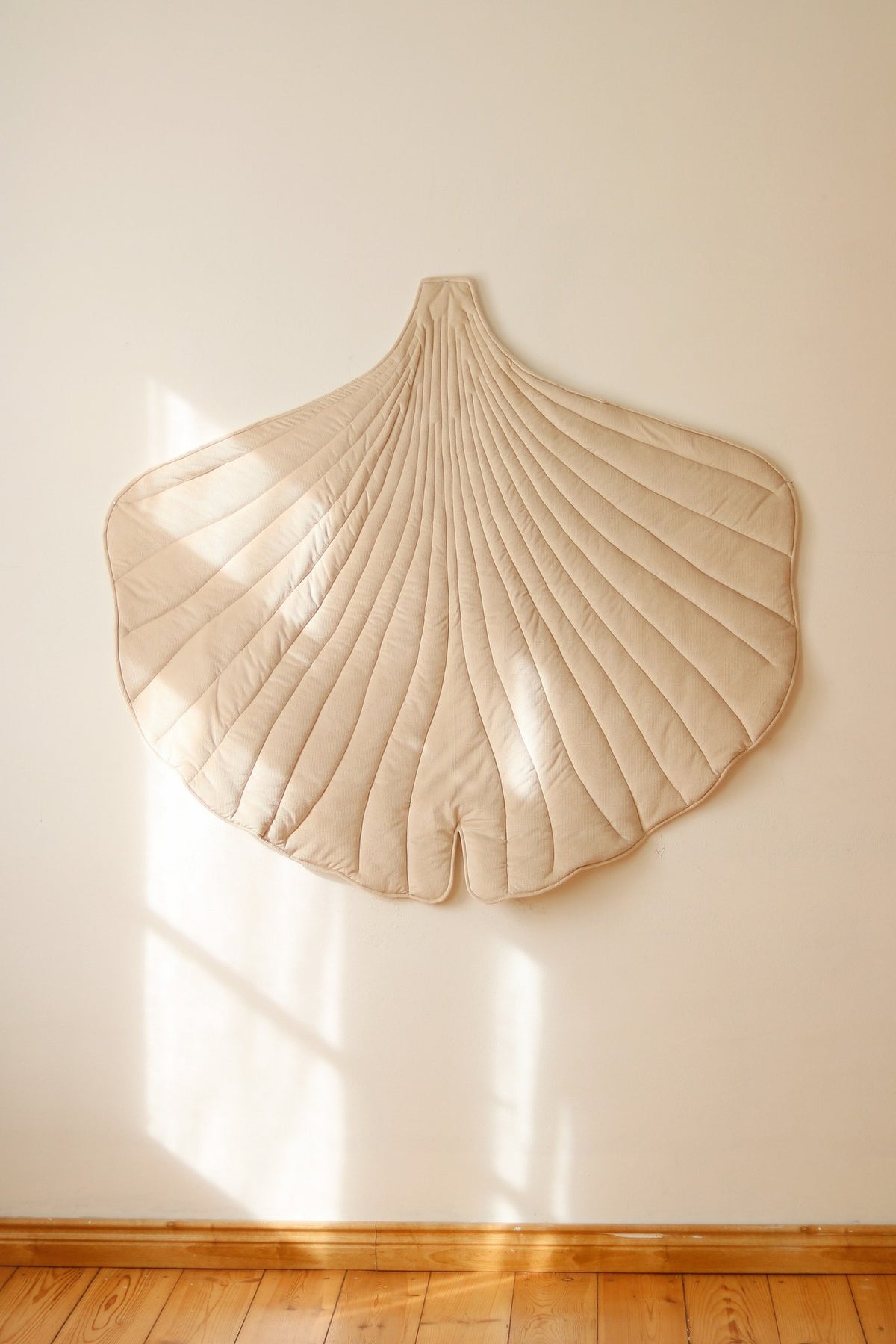 1. Cream ginkgo leaf playmat hanging on a white wall in a sunlit room, highlighting its quilted texture and organic shape