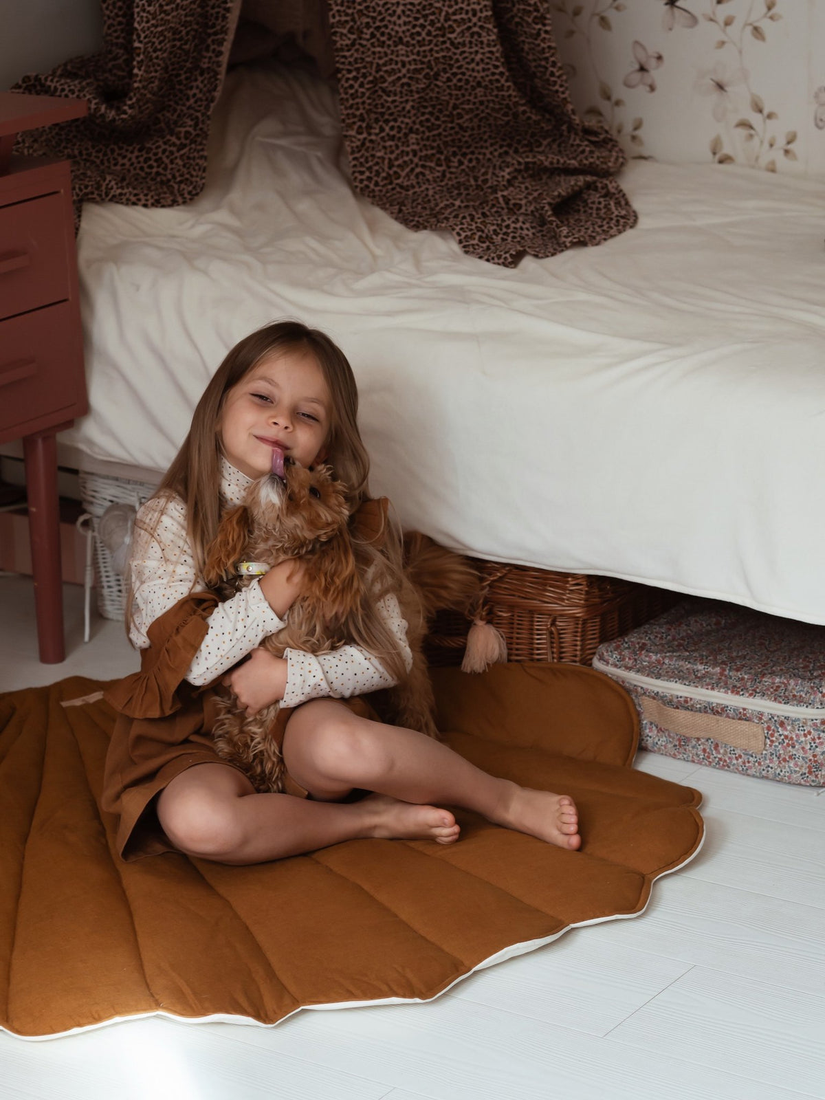 1. Young girl sitting on caramel shell playmat in a cozy bedroom setting, hugging a plush toy