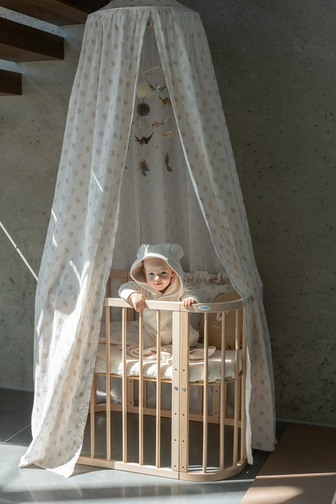 3. Baby in beige outfit inside wooden crib with floral canopy and hanging ornaments, natural light