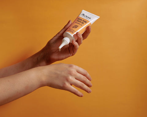 1. Person applying Nurme 100% Pure Shea Oil from twist cap tube onto hand against orange background