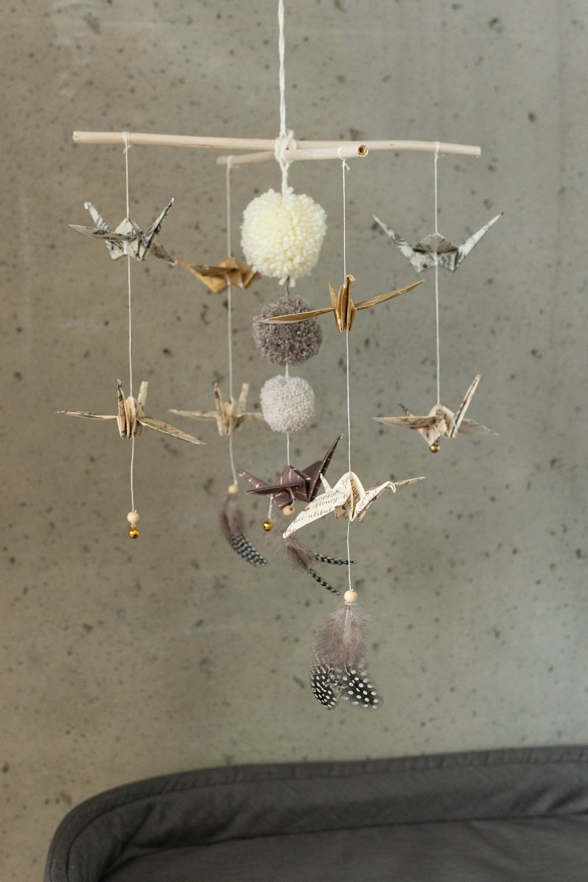 2. Moi Mili origami nursery mobile with gold and beige paper cranes, pompoms, and feathers hanging in front of concrete wall