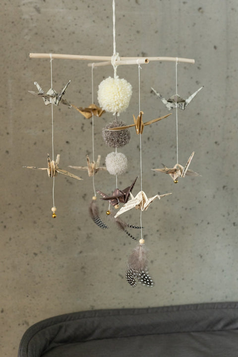 2. Moi Mili origami nursery mobile with gold and beige paper cranes, pompoms, and feathers hanging in front of concrete wall