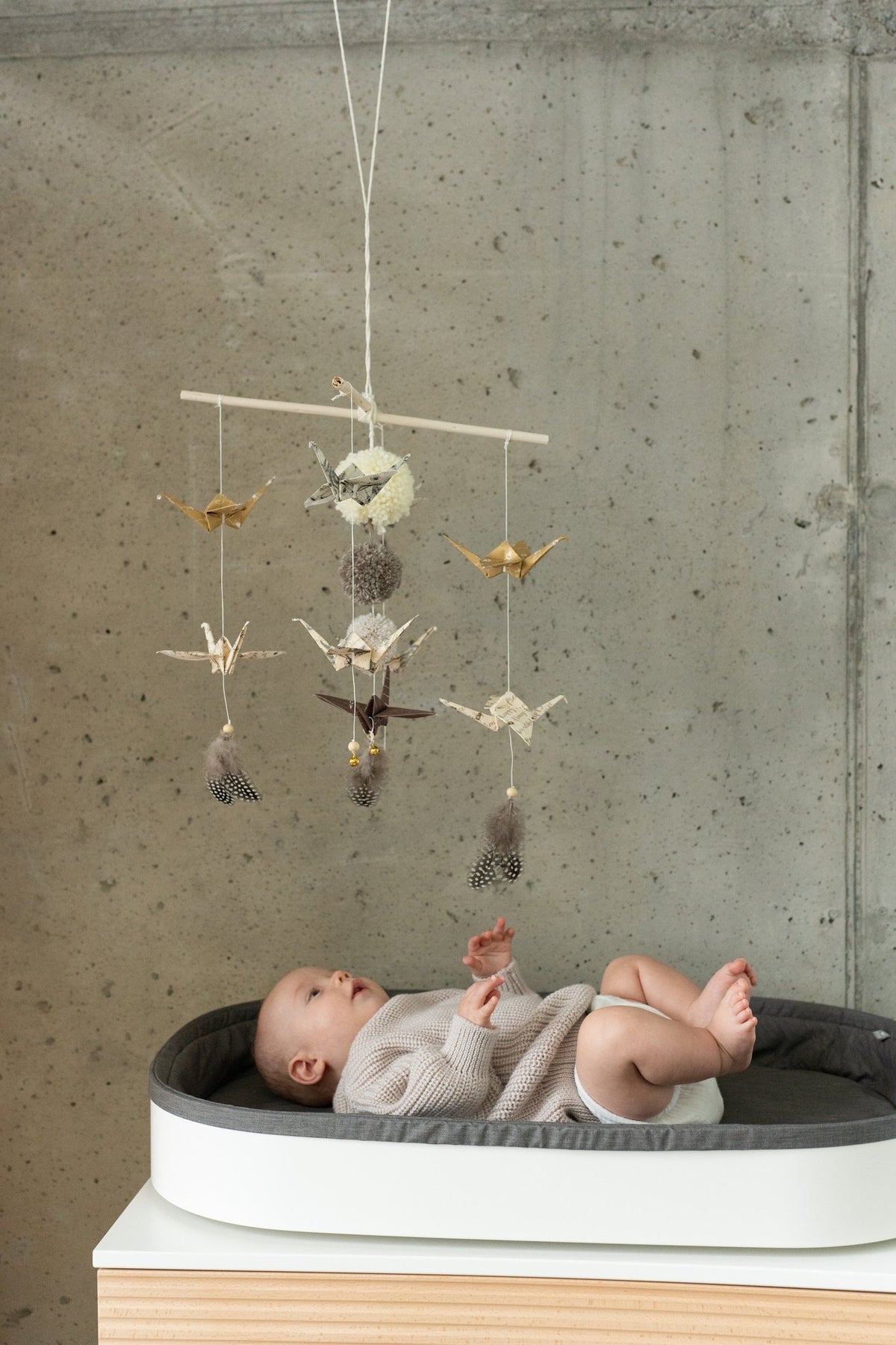 3. Baby in crib gazing at Moi Mili origami mobile with paper cranes and pompoms against concrete backdrop