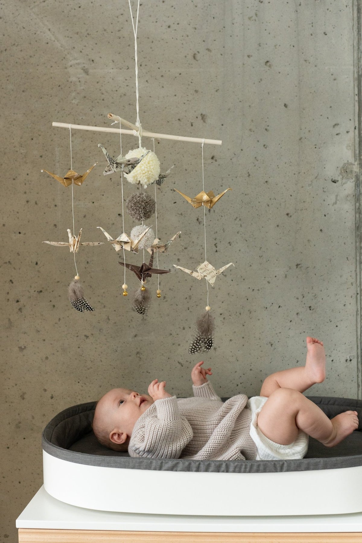 1. Baby lying in crib looking at Moi Mili origami nursery mobile with paper cranes and pompoms against concrete wall
