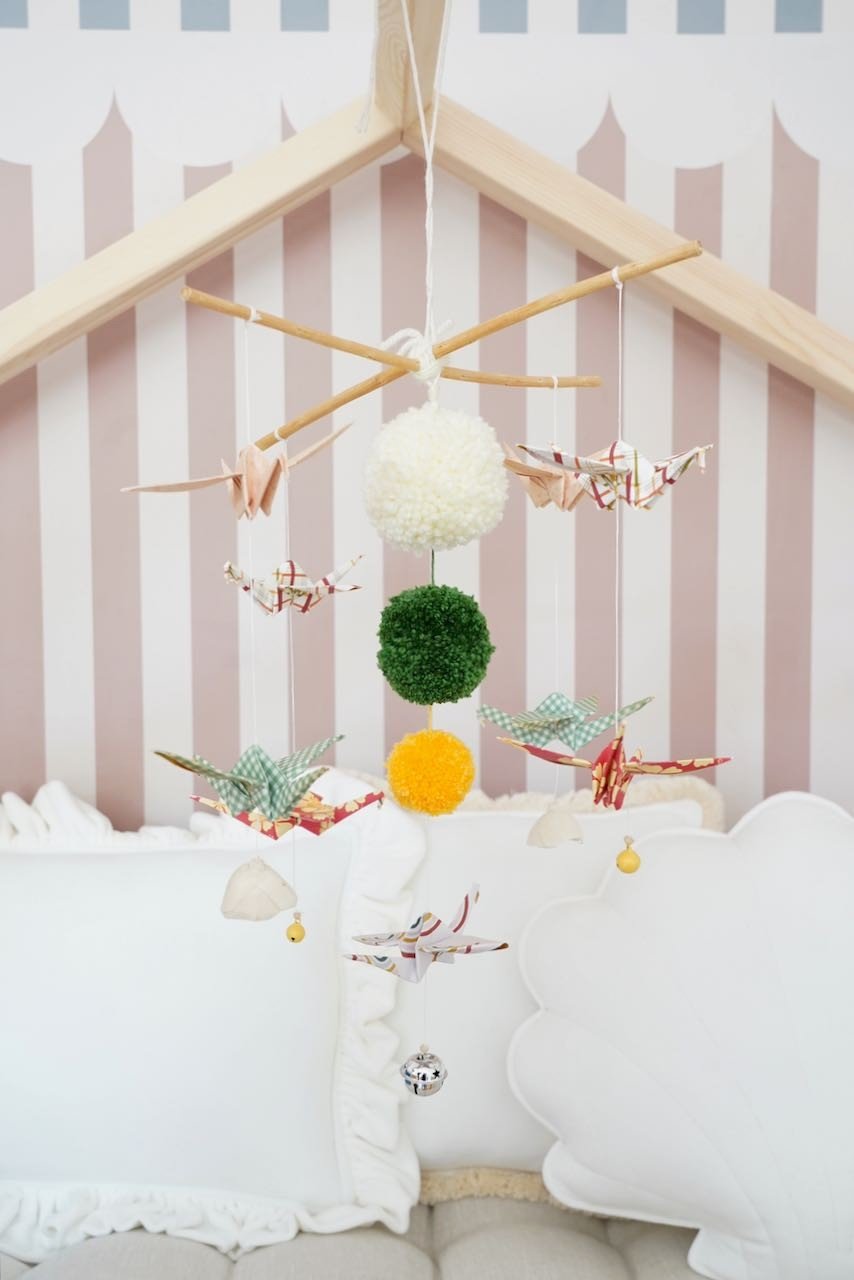 1. Handmade nursery mobile with origami cranes, yarn pompoms, and wooden sticks hanging above a crib in a cozy room with striped wallpaper
