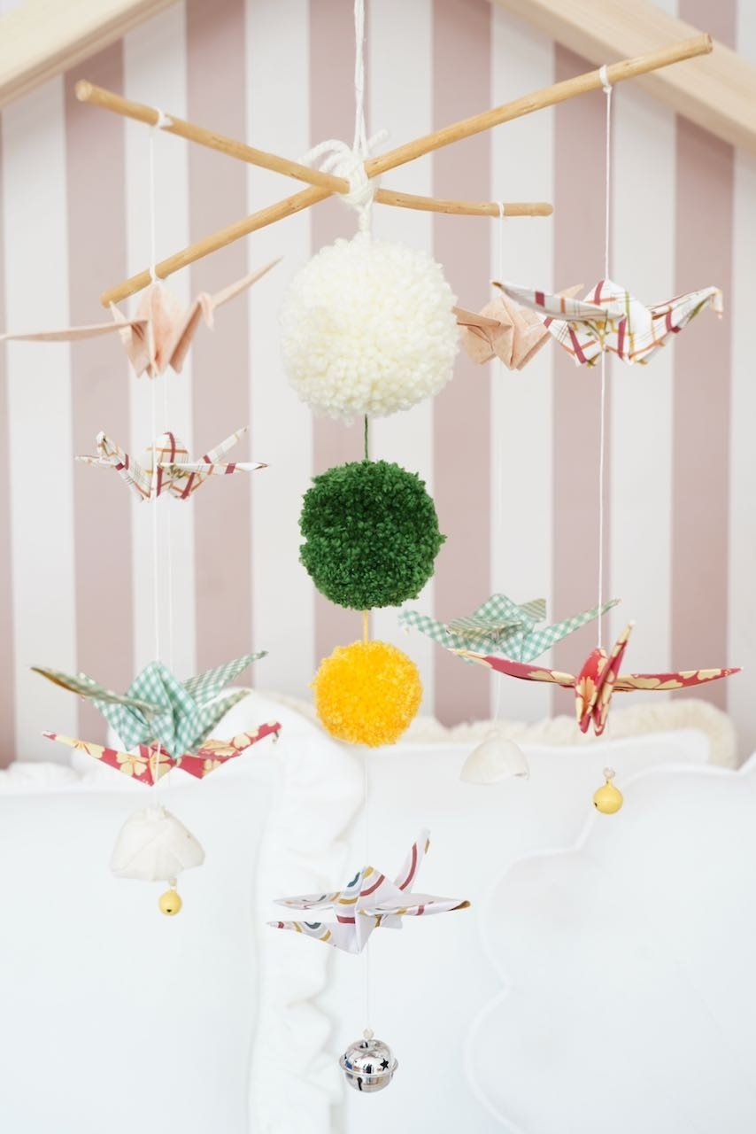 2. Nursery mobile with origami cranes and pompoms hanging in a cozy room, featuring a Scandinavian-inspired design with wooden sticks and bells