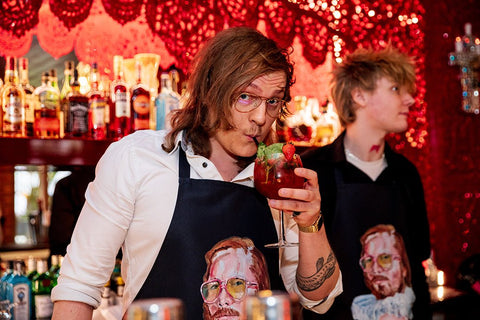 1. Two men wearing Katrin Valgemäe's 'Pervert in the Kitchen' aprons, one drinking a cocktail, in a vibrant bar setting