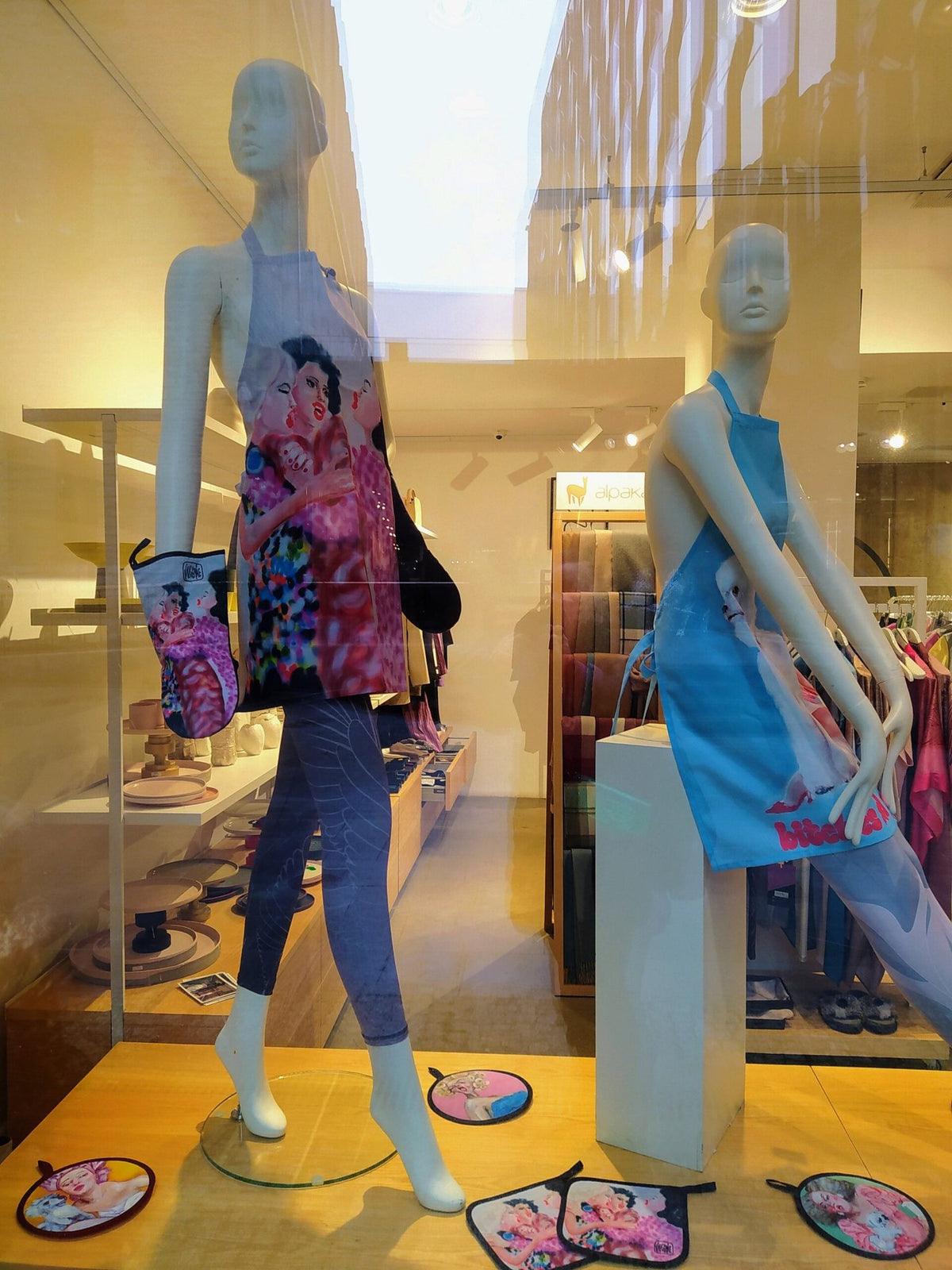 2. Store display with mannequins wearing Katrin Valgemäe aprons, one with a vibrant painting of women, set in a modern retail environment