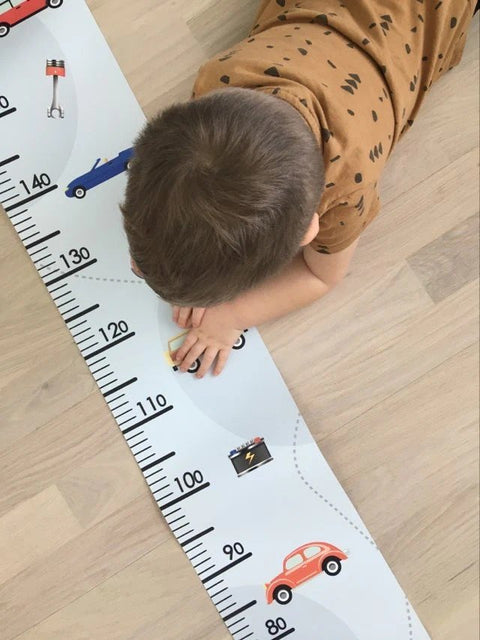 1. Child lying on floor interacting with retro cars-themed paper growth chart, featuring colorful car illustrations and measurement scale, on light wooden floor