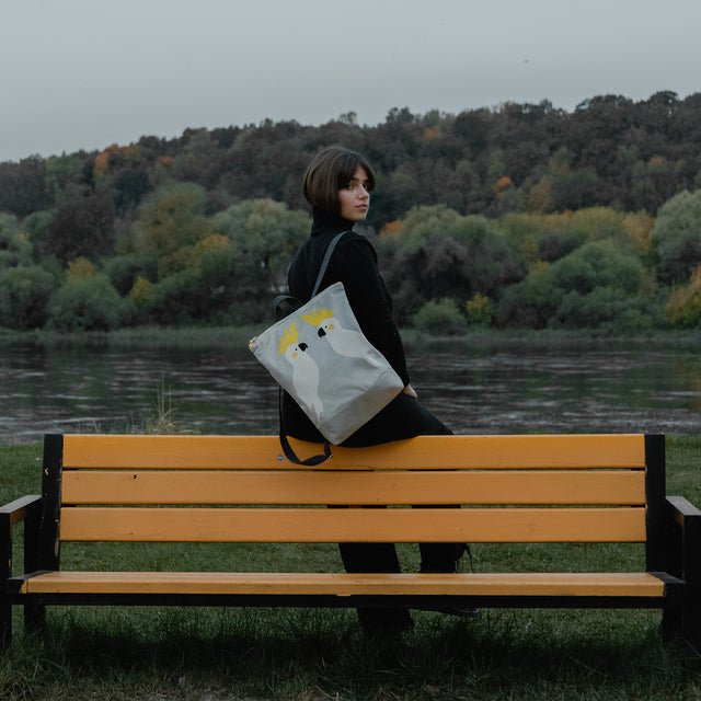 7. Woman on a bench with Muni Cockatoo backpack in grey velvet, featuring cockatoo design, with a scenic background