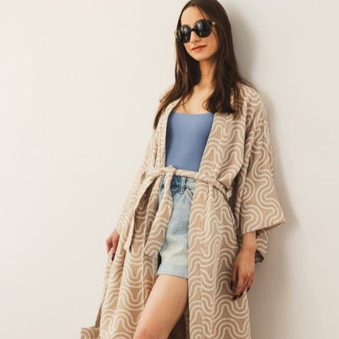 1. Woman wearing Patulove sand bamboo kimono with blue top and denim shorts, leaning against a white wall