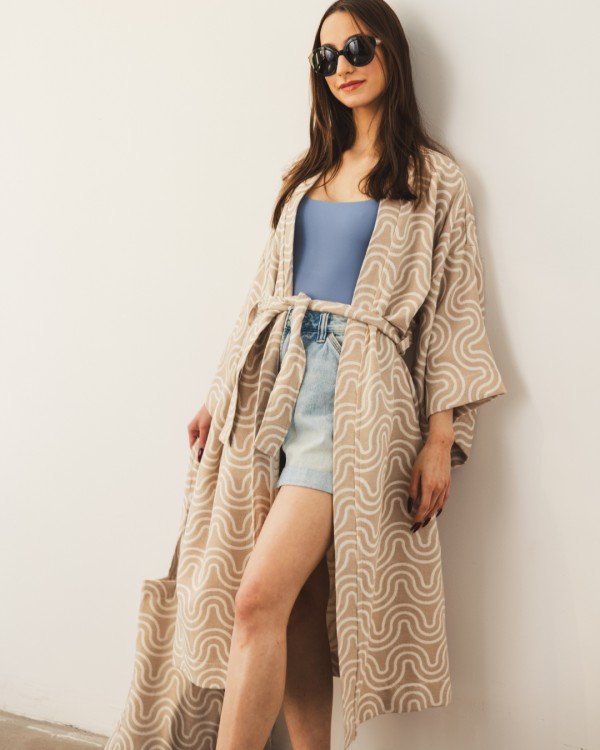 2. Woman in Patulove sand kimono with blue top and denim shorts, standing against a white wall