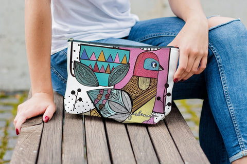 1. Woman sitting on a bench holding Garbanota makeup bag with Little Bird print, showcasing colorful artistic design