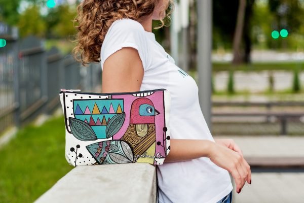 2. Woman leaning on railing with Garbanota Little Bird makeup pouch, highlighting vibrant illustration and casual style