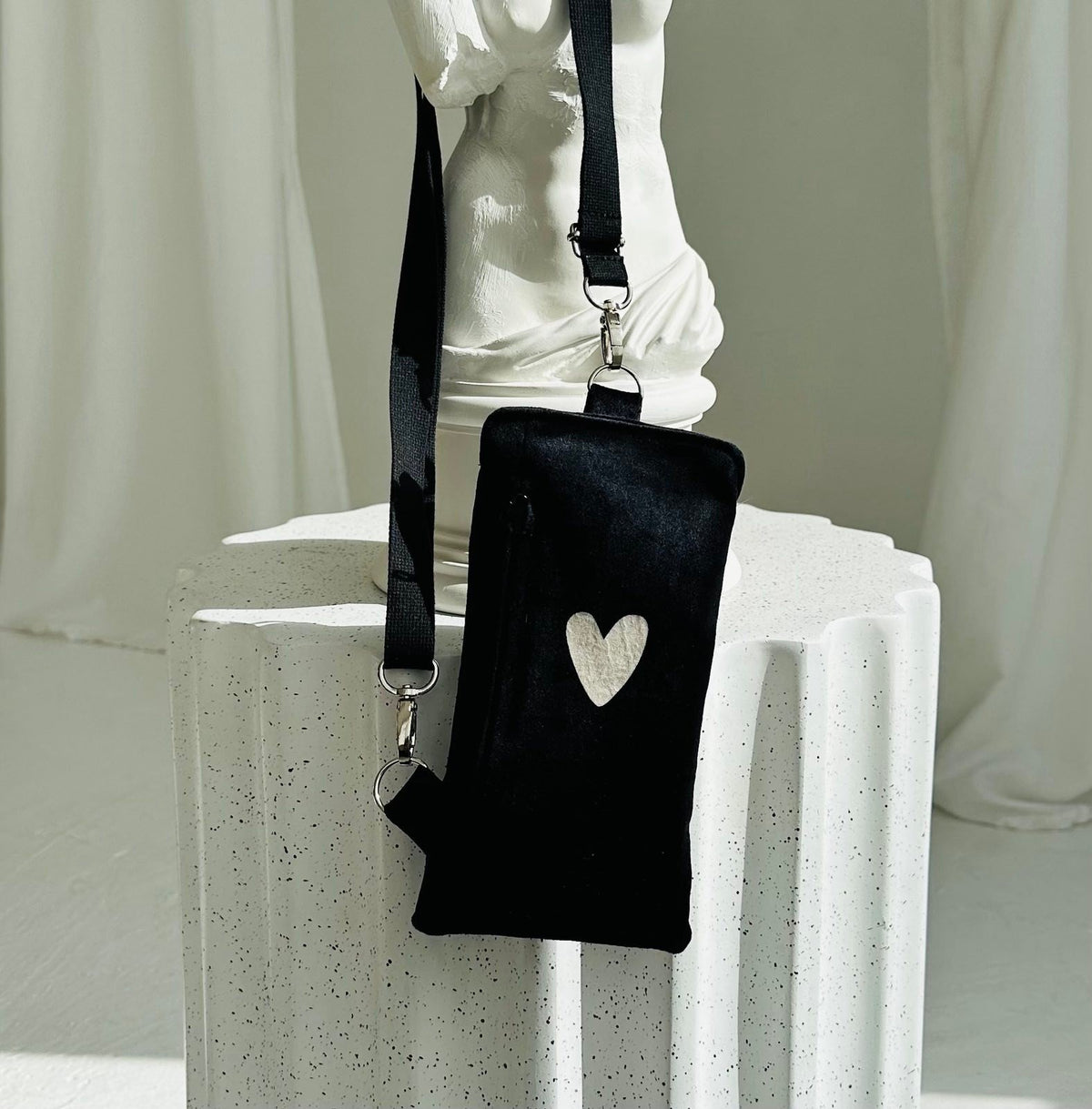 2. Black velour phone bag with heart design by Garbanota, displayed on a white pedestal with adjustable strap