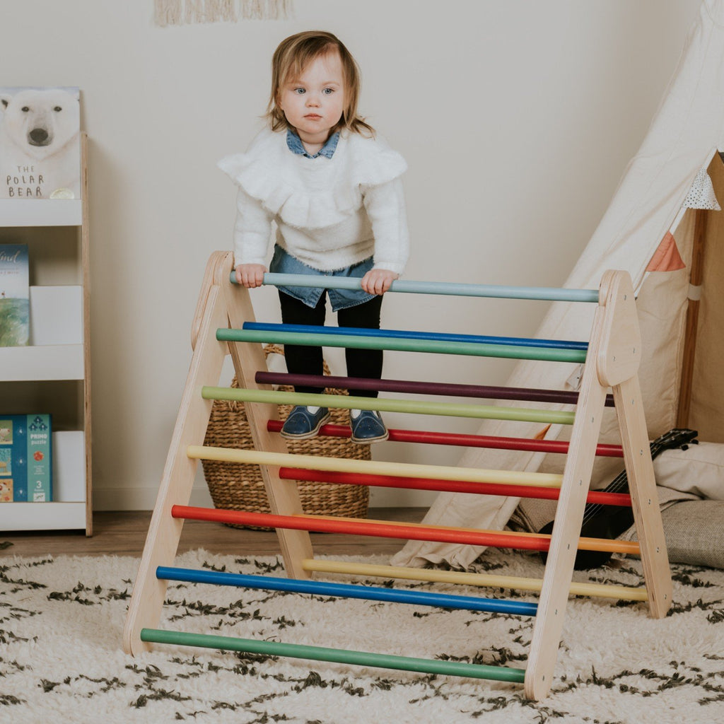 1. Child standing on leg&go Pikler Triangle with rainbow bars in playroom with books and teepee
