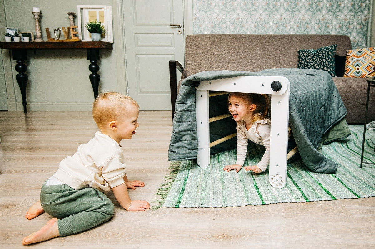 3. Two toddlers playing with a white Pikler triangle covered with a blanket in a cozy living room