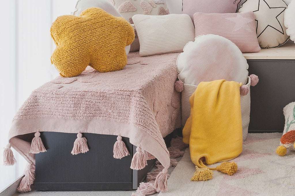 2. Mustard cloud pillow on pink tasseled bedspread with various pillows and a yellow blanket in a children's room