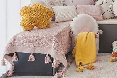2. Mustard cloud pillow on pink tasseled bedspread with various pillows and a yellow blanket in a children's room