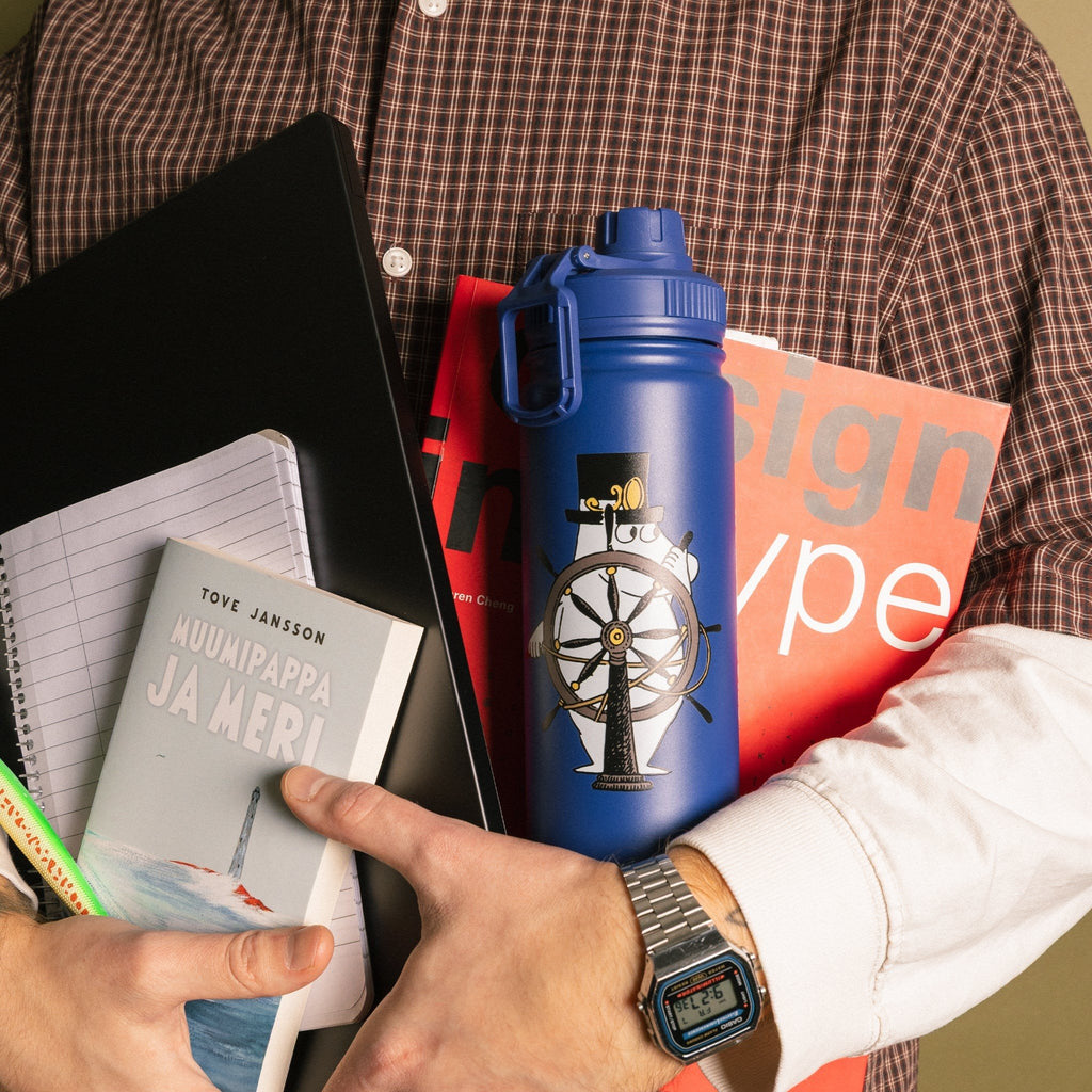 1. Person holding navy Moominpappa thermal bottle with Moomin design, books, and magazines in casual setting