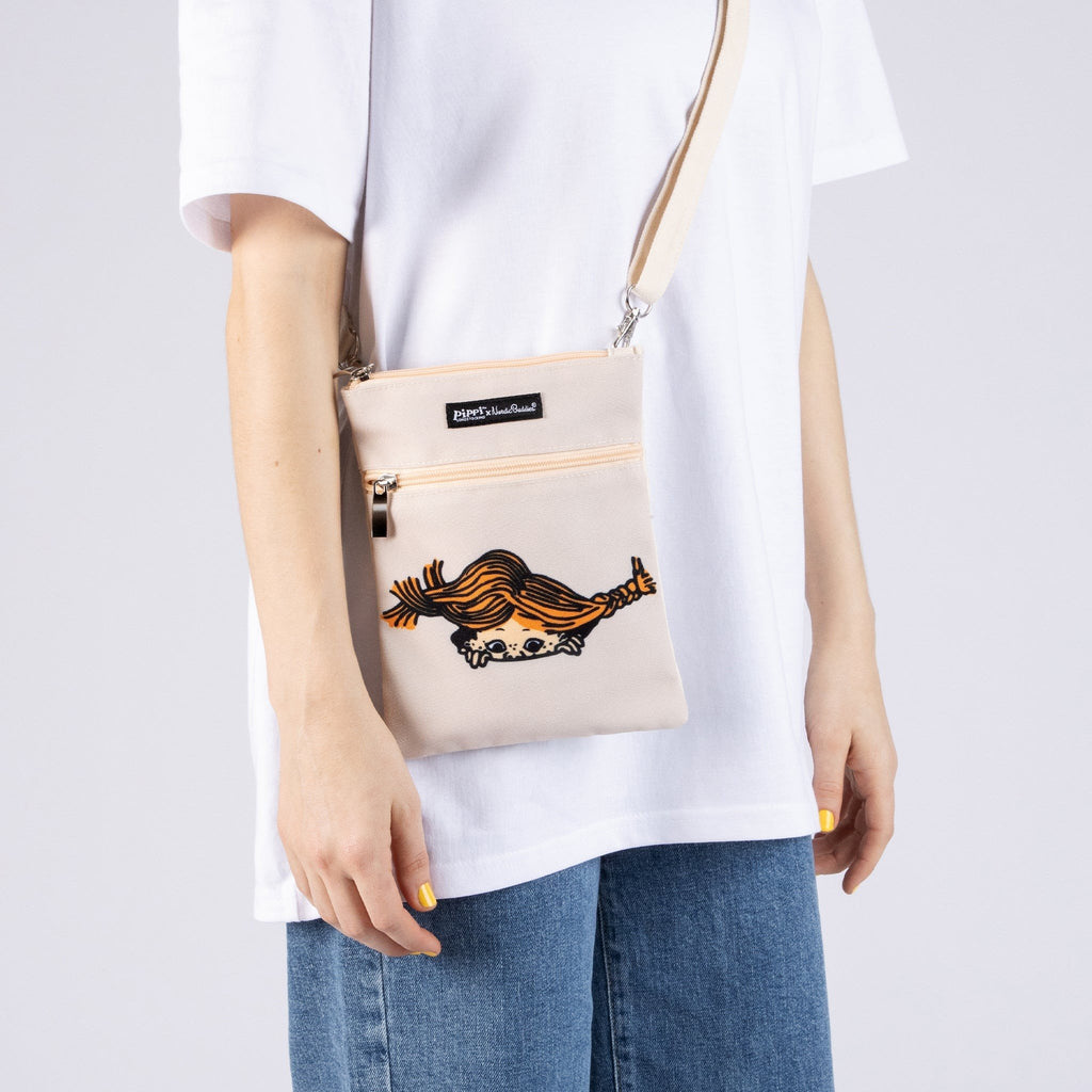 1. Women's Pippi Passport Bag in beige by Moomin, worn crossbody with casual outfit in studio setting