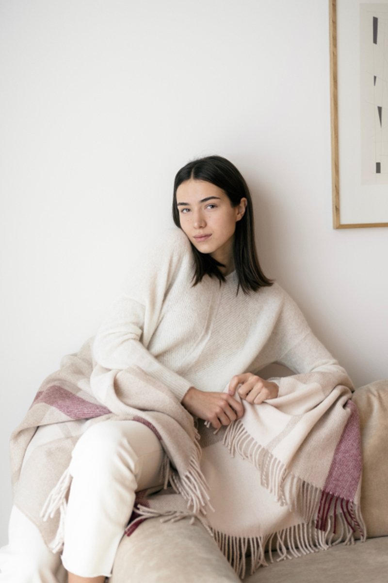 1. Woman wrapped in Alpaka Natural Alpaca Wool Throw in Rosé/Beige, sitting on a couch in a cozy setting