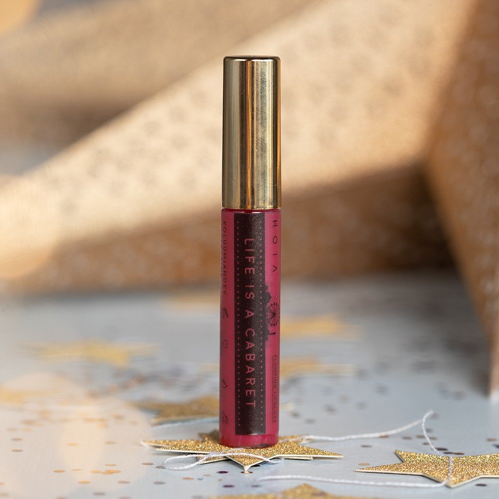 1. HOIA homespa plumping lip gloss in deep red with gold cap, labeled 'Life Is A Cabaret', standing on decorative star background, designed to volumize and moisturize lips with plant extracts.