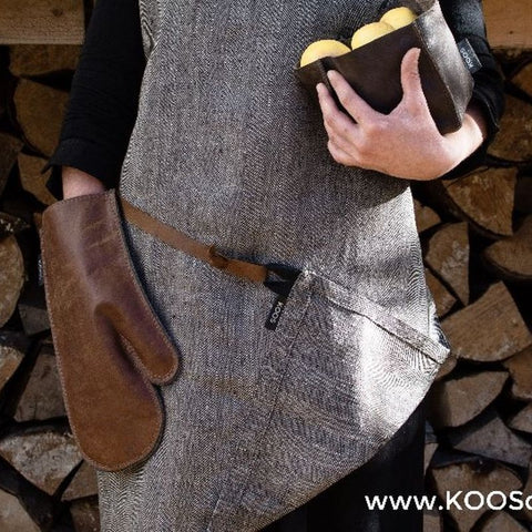2. Close-up of KOOSdisain herringbone linen apron with leather details, showing pocket with lemons and brown oven mitt against a woodpile background