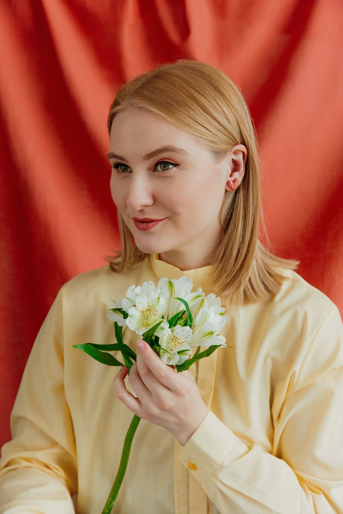 1. Woman wearing Rozenthal pink dotted porcelain heart studs with 24k gold accents, holding flowers against a red background