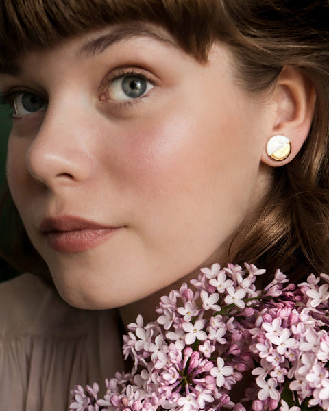 1. Woman wearing Rozenthal porcelain round stud earrings in white and gold, shown in lifestyle setting with flowers