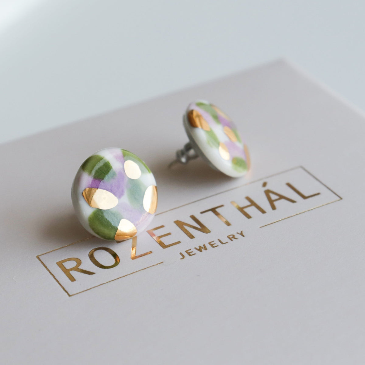4. Rozenthal Porcelain Tache Studs in green and violet on branded packaging, highlighting 24k gold accents