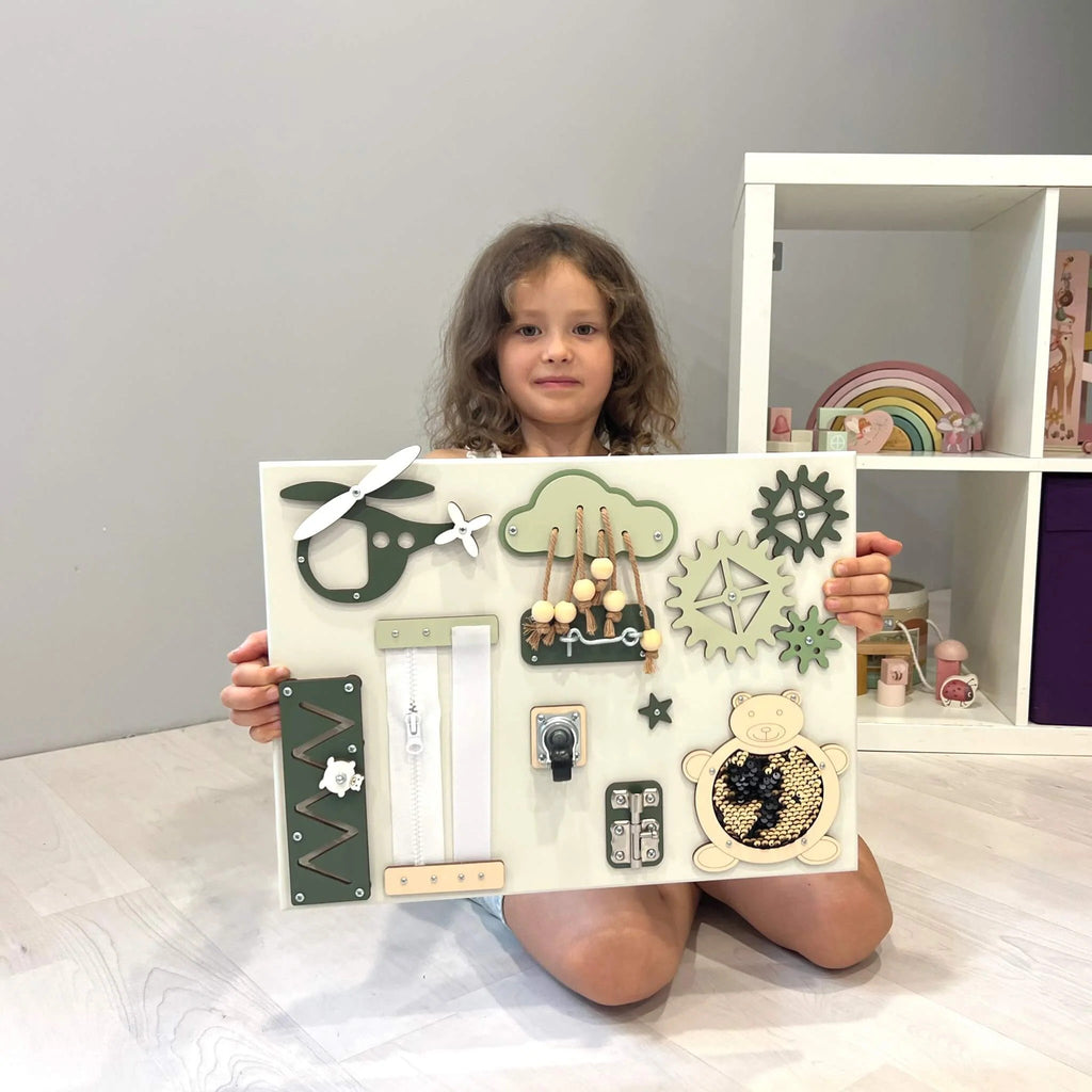 1. Child holding a green and beige baby sensory busy board with various interactive elements in a playroom setting