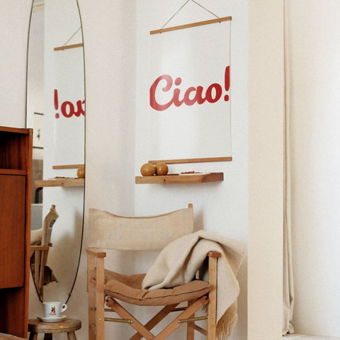 3. 'Ciao!' poster in red typography reflected in a mirror, adding elegance to a cozy room with a wooden chair and decorative shelf