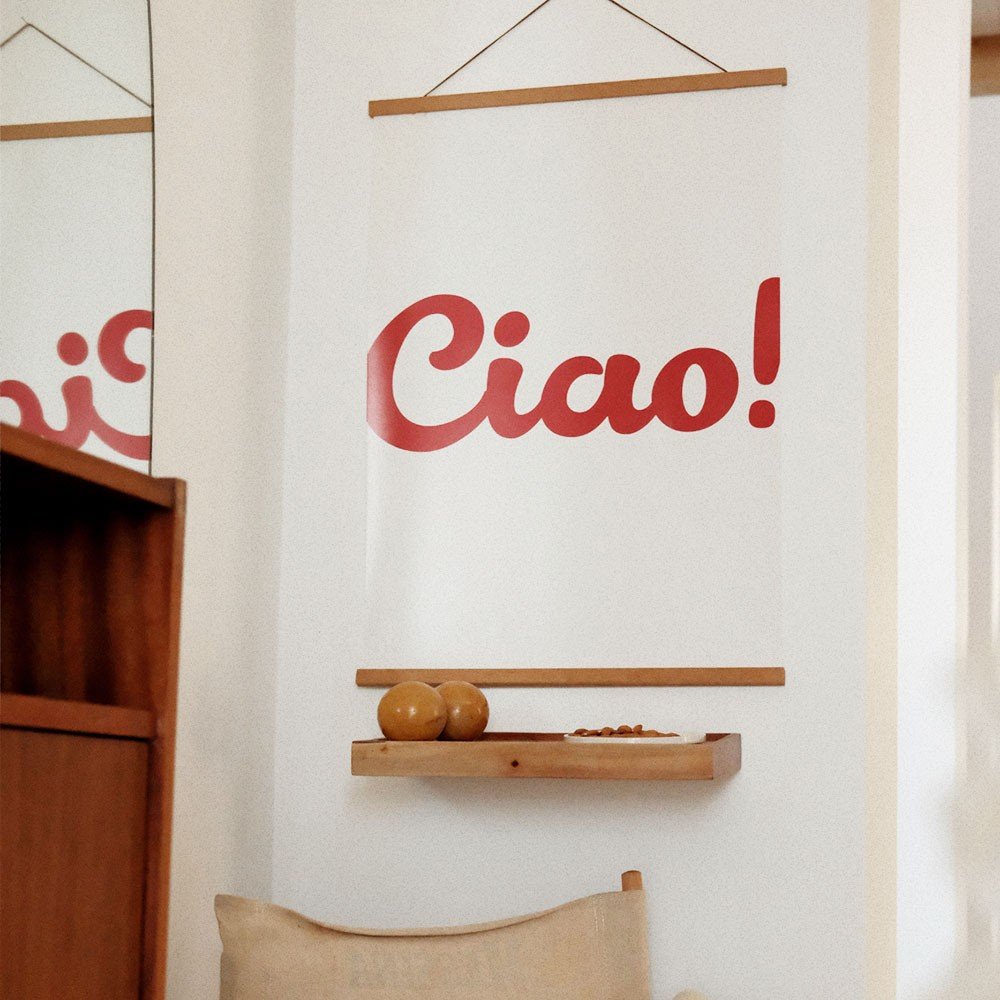 1. Minimalist 'Ciao!' poster in red typography hanging on a white wall above a wooden shelf with decorative items, enhancing a cozy interior setting
