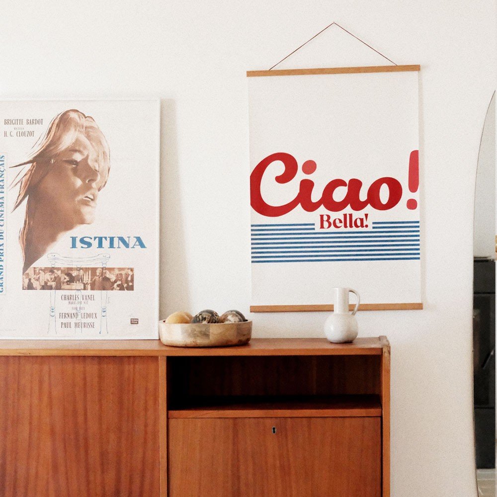 5. 'Ciao Bella' poster with red and blue design hanging next to a vintage poster on a wooden cabinet