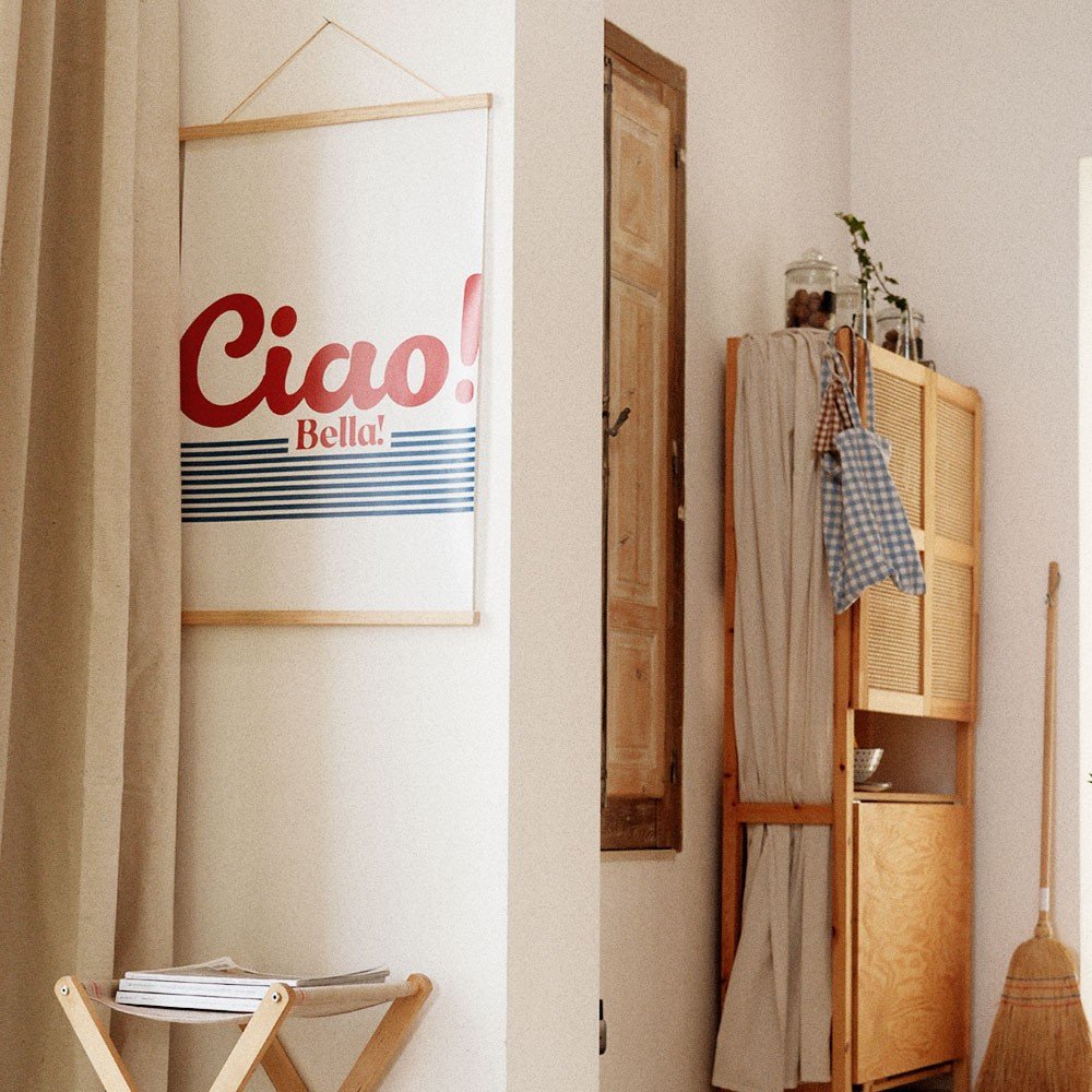 1. 'Ciao Bella' poster with red typography and blue stripes hanging in a cozy room with wooden furniture and neutral curtains