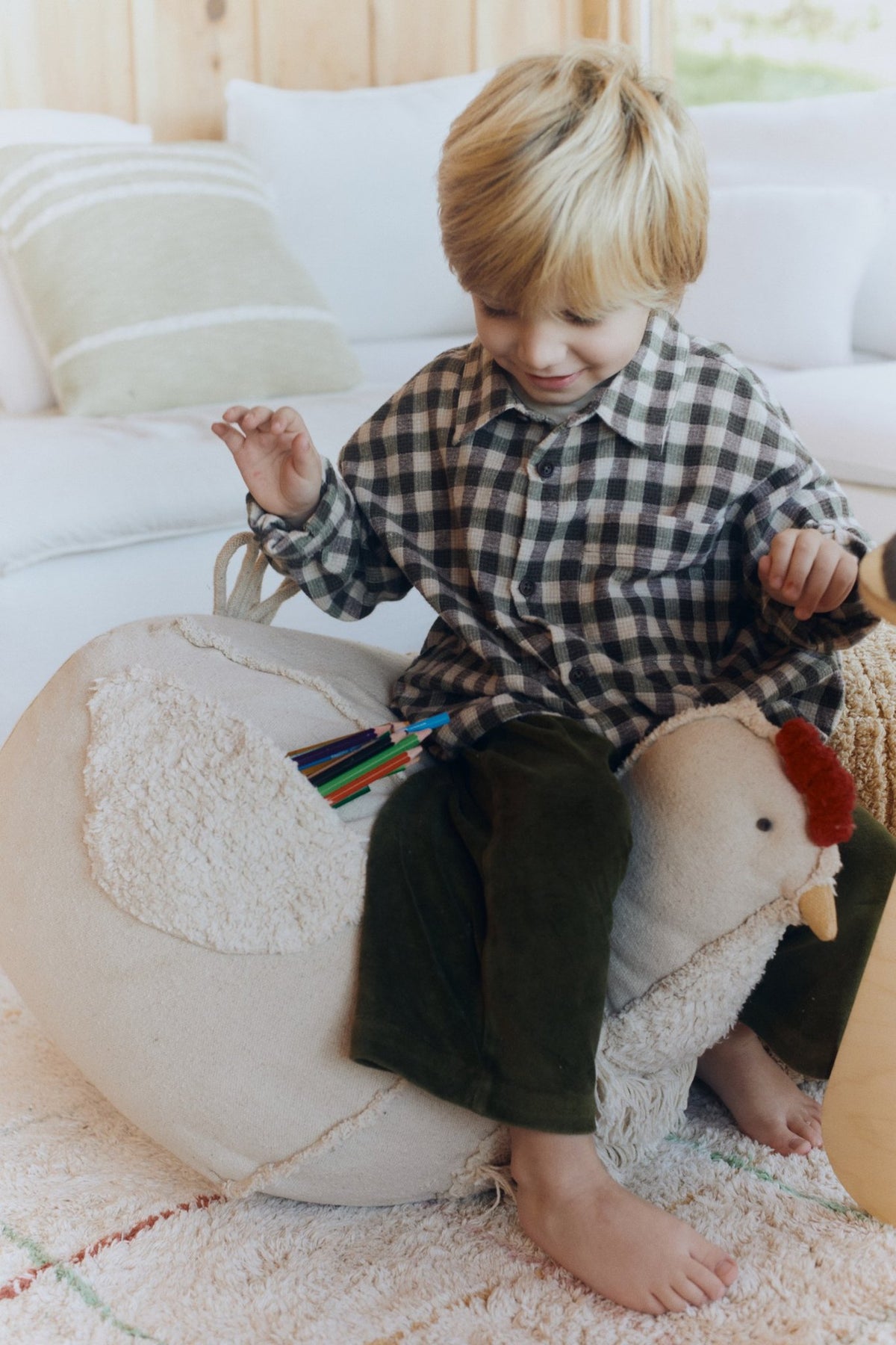 1. Child sitting on beige chicken pouf in cozy living room, holding colored pencils