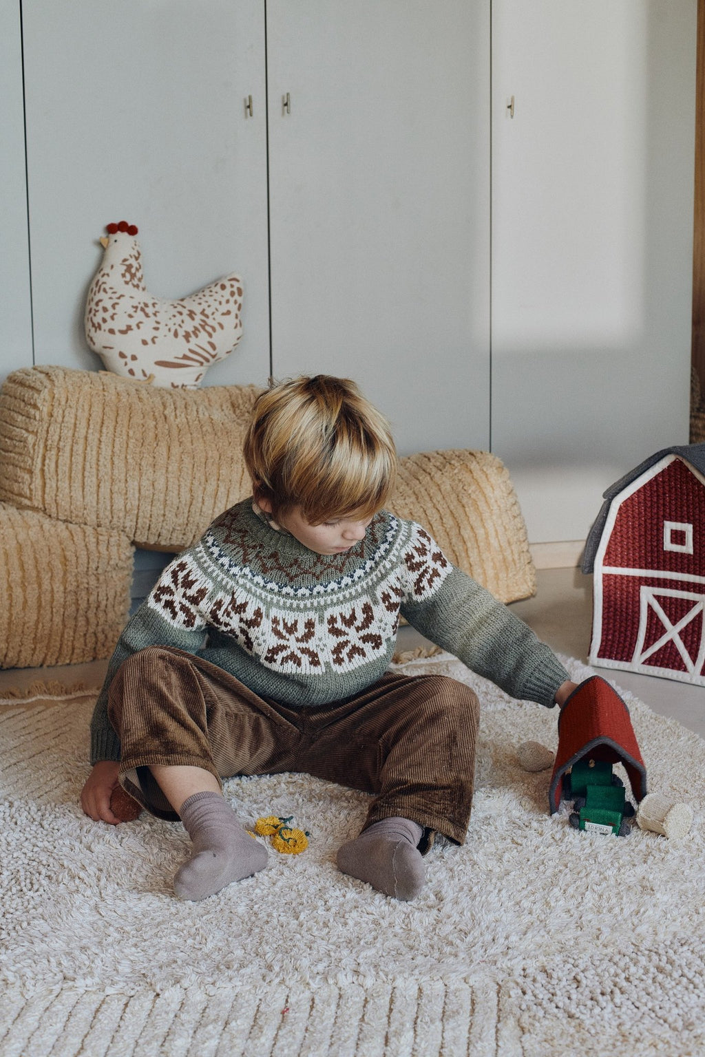 1. Child playing with toys on Lorena Canals Pouf Haystack in light honey, set in a cozy room with a toy barn and decorative elements