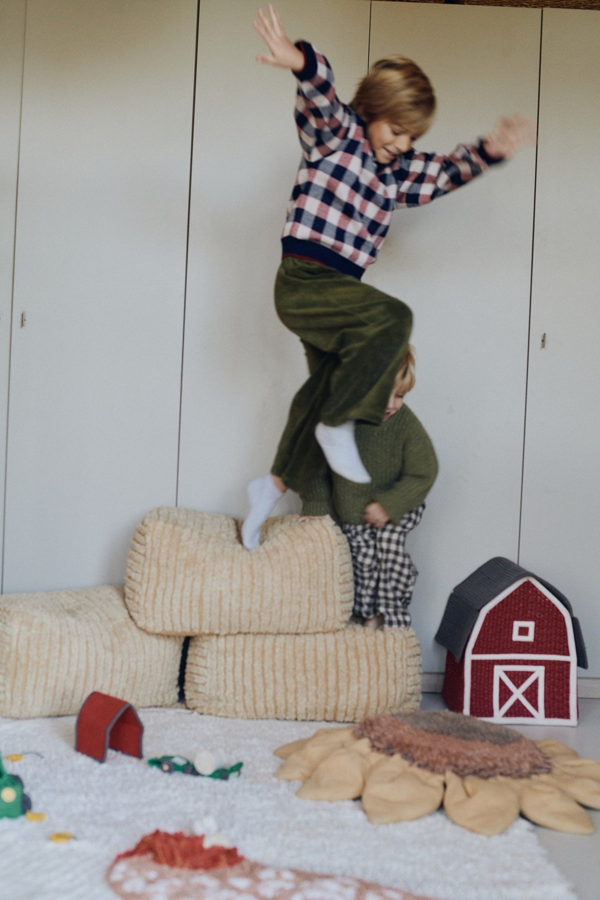 1. Two children playing on stacked Lorena Canals Pouf Haystacks in light honey, in a playful, rustic-themed room with a toy barn