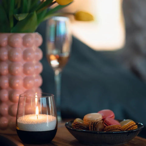 1. Lit candle in glass holder on table with pink vase, champagne glass, and macarons in cozy setting
