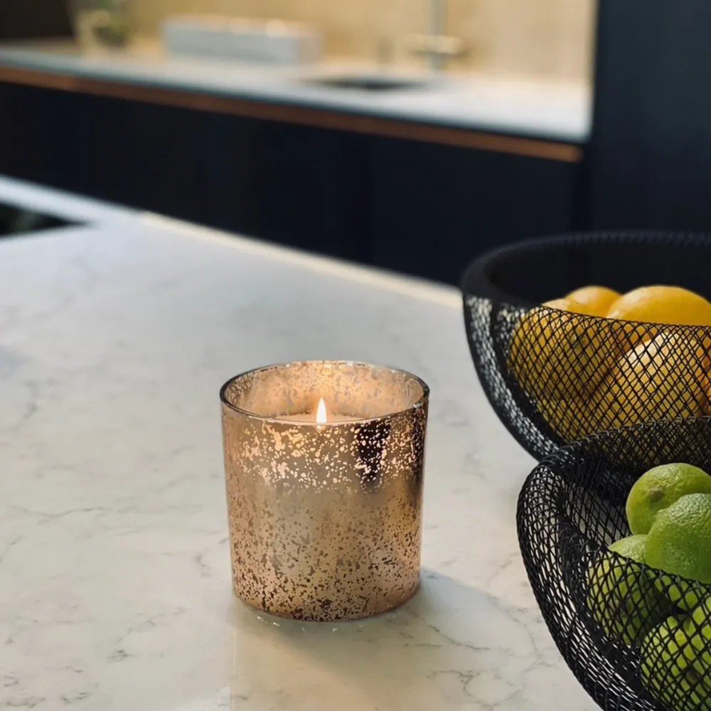 1. Lit candle in a decorative glass holder on a kitchen counter, next to a fruit basket, highlighting the ambiance of the powdered candle refill kit