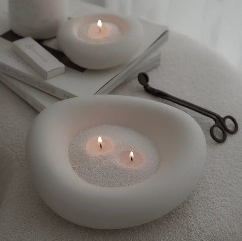 1. Two white ceramic bowls filled with lit candle sand, placed on books and a soft surface, with candle wick trimmer nearby