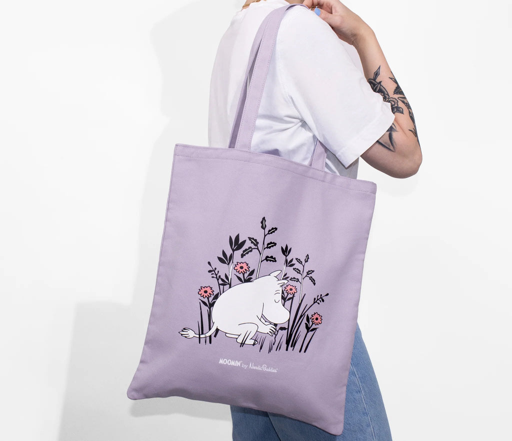 1. Woman carrying Moomintroll tote bag in vibrant purple, featuring floral design, in lifestyle setting