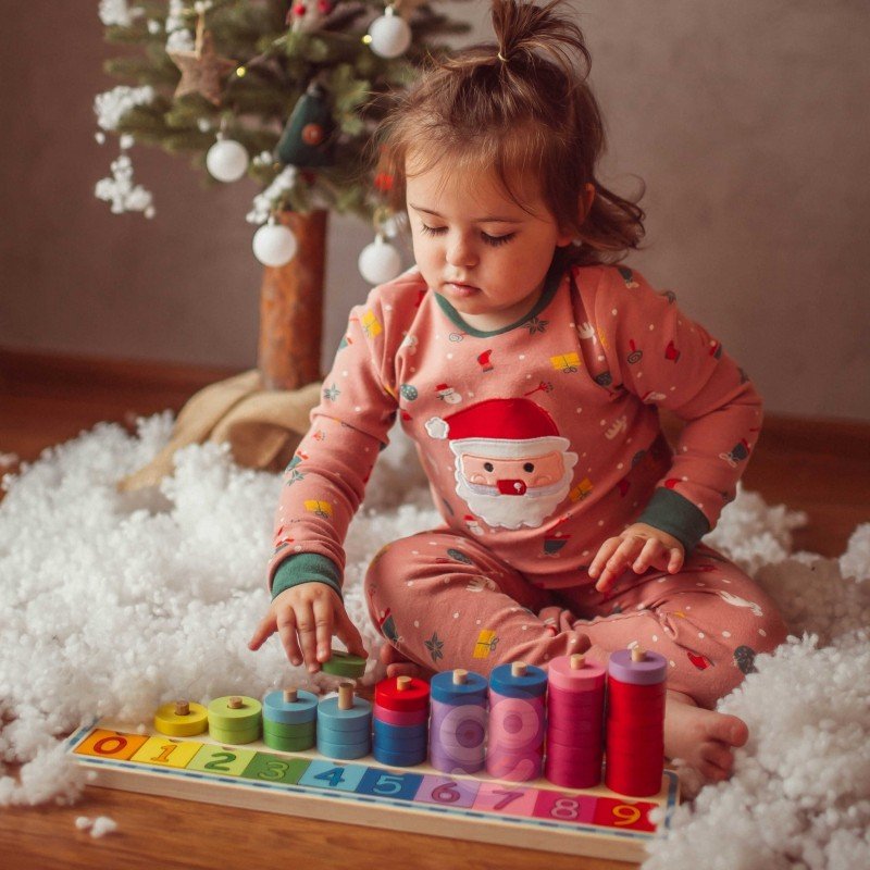 5. Child in Santa-themed pajamas playing with colorful wooden counting puzzle under a Christmas tree