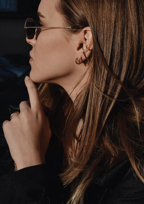 1. Woman wearing NO MORE Radius gold earrings with sunglasses, styled in an outdoor setting