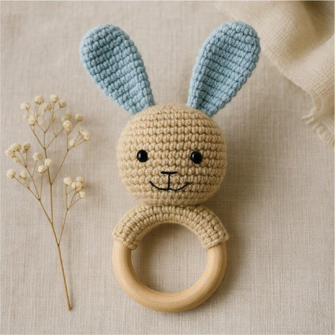 1. Handmade crocheted bunny rattle with blue ears and natural wooden ring on beige fabric background, ideal for teething and soothing, crafted by Beebila in Estonia