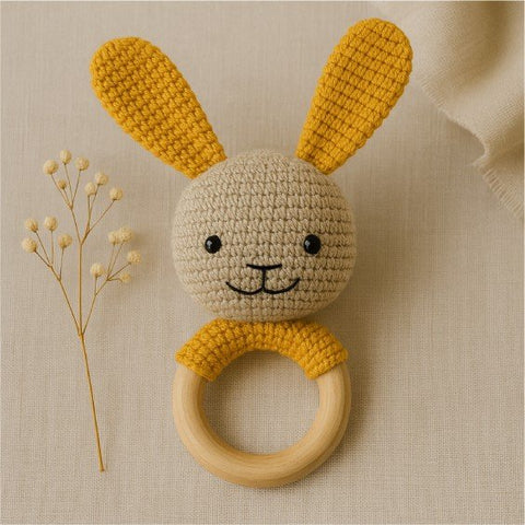1. Handmade yellow crocheted bunny rattle with natural wooden ring on beige fabric background, featuring safety eyes and hand-stitched details, ideal for teething and soothing gums.
